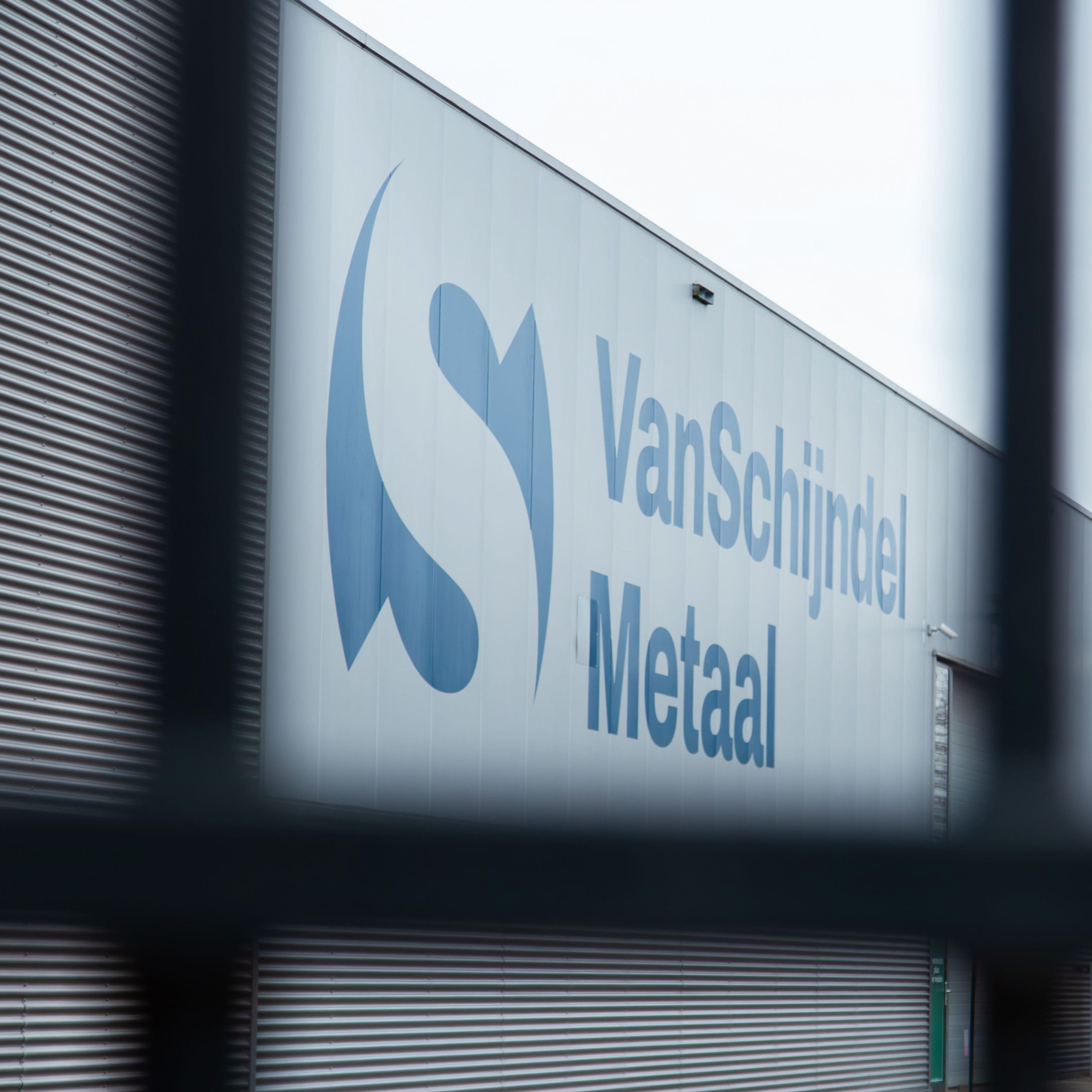 Logo van Van Schijndel Metaal zichtbaar door hekwerk bij bedrijfspand